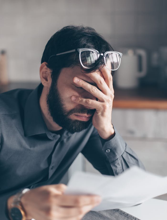 Man with glasses on head holds head in frustration while reading financial documents, indicating stress.